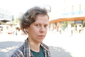 Mari med litet leende, en solig dag på torget i Farsta centrum. En sommarbris fläktar i håret. 